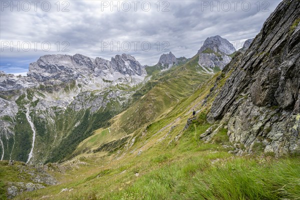Mountaineer on a narrow hiking trail on a steep meadow slope, descent from the Raudenspitze, view into the Val Fleons valley with rocky mountain peaks, Carnic Main Ridge, Carnic Alps, Carinthia, Austria, Europe