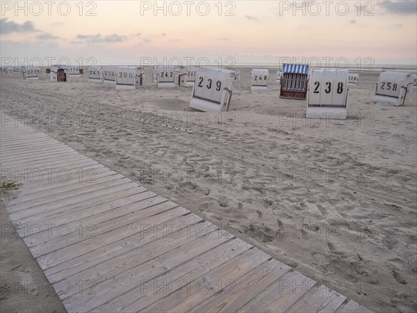 A wooden path leads to empty beach chairs on a quiet beach under a cloudy evening sky, setting sun on a beach with beach chairs and clouds in the sky, spiekeroog, germany