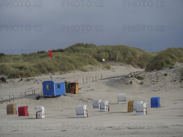 Several beach chairs are standing on the sandy beach in front of a hilly dune landscape and a cloudy sky, colourful beach chairs on the beach and in the dunes under an overcast sky, red wooden boat and yellow beach chair, Juist, Germany, Europe