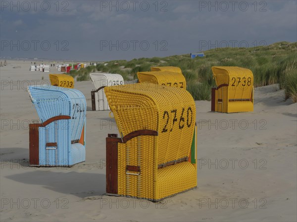 View of colourful beach chairs in front of cloudy sky, some baskets in the sand with dune grass, colourful beach chairs on the beach and in the dunes with cloudy sky, red wooden boat and yellow beach chair, Juist, Germany, Europe