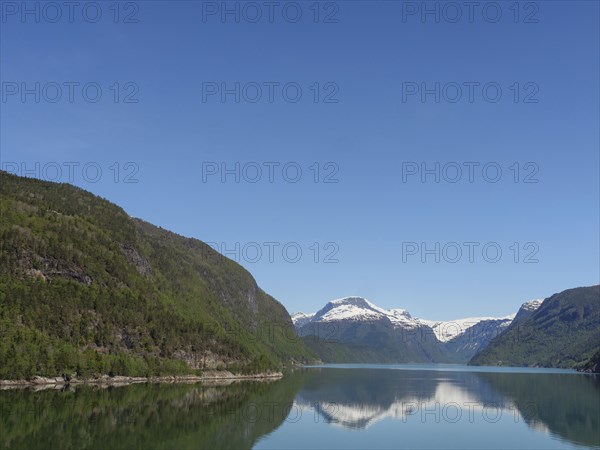 A calm lake, surrounded by snow-capped and forested mountains, under a clear blue sky, calm water in a fjord with snow-capped mountains and green slopes with trees, Eidjord, Norway, Europe