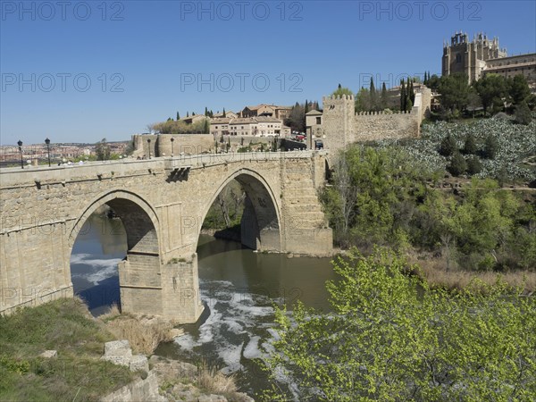 Stone bridge over the river, old city with cathedral in the background, many trees and blue sky, historic stone bridge with an old tower and an old city in the background over a wild river, Toledo, Spain, Europe