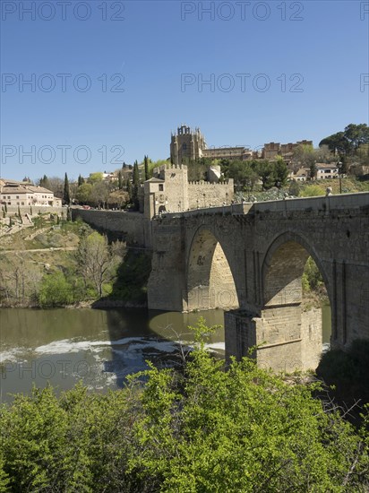 Stone bridge with arches over a river, surrounded by a cityscape and vegetation, sunny weather, historic stone bridge with an old tower and an old town in the background over a wild river, Toledo, Spain, Europe