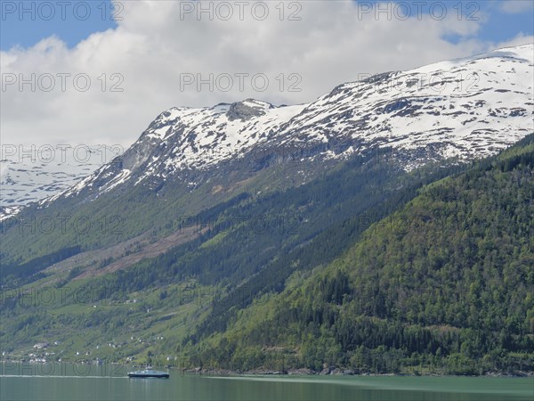Snow-capped mountain peaks rising into the blue sky with a calm body of water and green mountainsides, shimmering green water in a fjord with snow-capped mountains and green trees, eidfjord, Norway, Europe