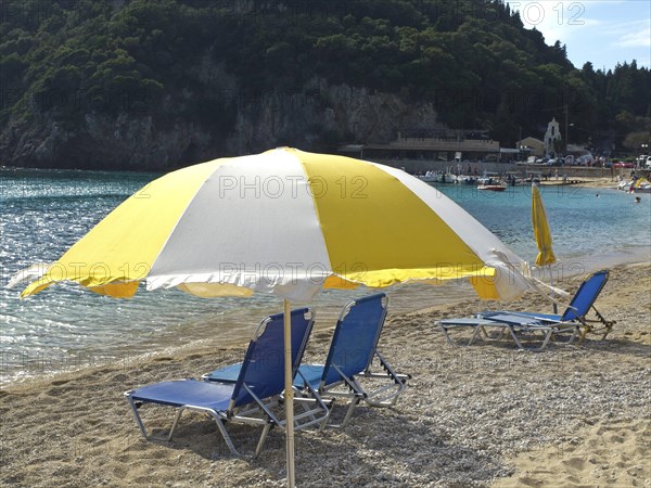 Yellow and white parasols and blue deckchairs on the beach with coast in the background, parasols and deckchairs on a small beach with green blue water, corfu, greece
