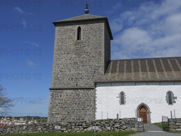 Stone church with a stone wall in the foreground and a clear blue sky, old stone church and many gravestones by the water with ships in the background, haugesund, norway