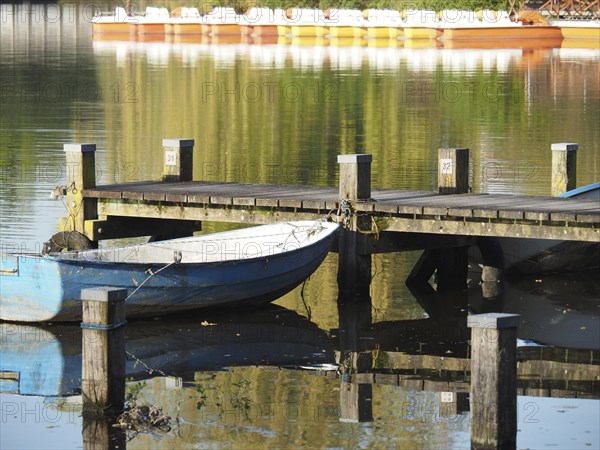 A boat is moored next to a jetty that is reflected in the water, surrounded by peaceful nature, rowing boats and pedal boats on a small wooden jetty in the lake surrounded by trees, Borken Germany
