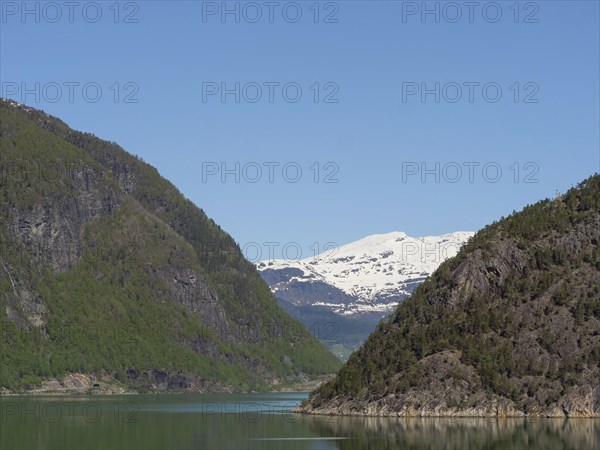 A narrow lake between large rocky mountains, with snow-capped peaks in the background and a blue sky, calm water in a fjord with snow-capped mountains and green slopes with trees, Eidjord, Norway, Europe