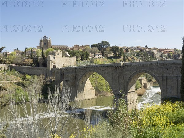 Historic stone bridge with arches over a river, surrounded by vegetation and cityscape, sunny weather, historic stone bridge with an old tower and an old town in the background over a wild river, Toledo, Spain, Europe