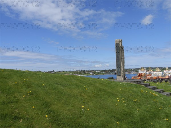 Green meadow with a monument on a hill under a blue sky, old stone church and many gravestones on the waterfront with ships in the background, haugesund, norway