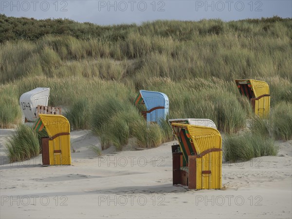 Beach chairs in different colours stand along the dunes, protected by high grasses, colourful beach chairs on the beach and in the dunes in cloudy weather, red wooden boat and yellow beach chair, Juist, Germany, Europe