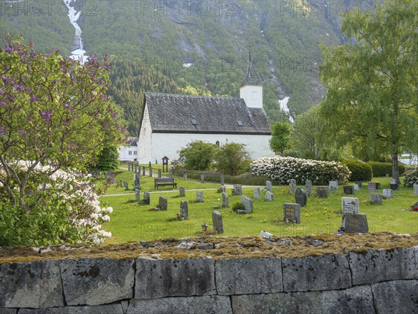 Cemetery with church and gravestones, surrounded by green landscape and high mountains, gravestones with a small white church in front of a mountain panorama in spring, eidfjord, norway