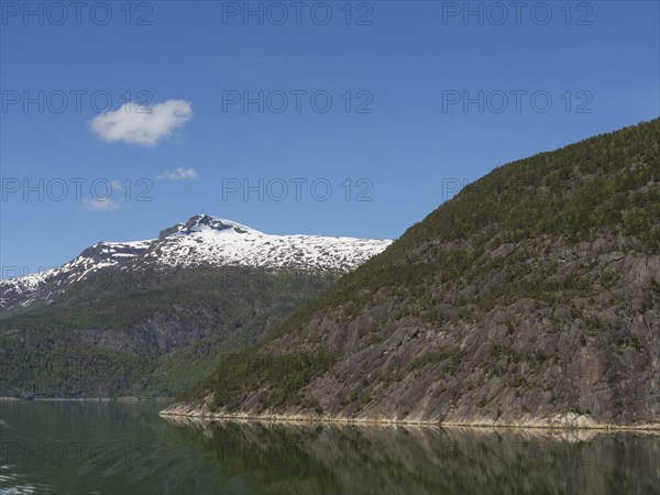 A calm lake surrounded by high green mountains with snow-capped peaks under a clear blue sky, calm water in a fjord with snow-capped mountains and green slopes with trees, Eidjord, Norway, Europe
