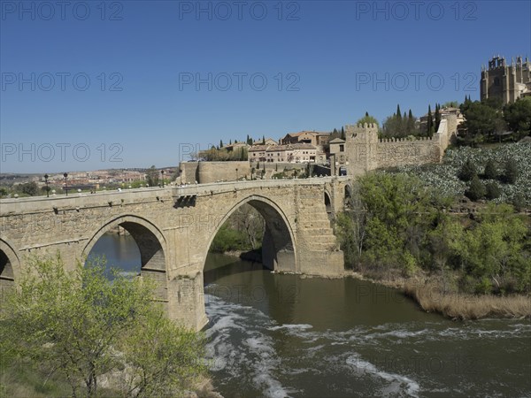 Stone bridge over a river with historic buildings and vegetation under blue sky, historic stone bridge with an old tower and an old town in the background over a wild river, Toledo, Spain, Europe