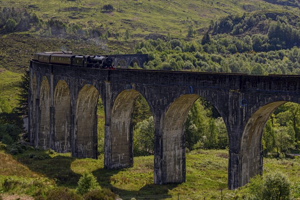 An old train crosses an impressive stone viaduct bridge stretching over a wooded glen under a clear sky, Scotland, United Kingdom, Europe