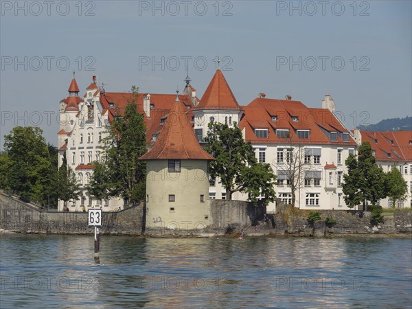 A castle with a red roof on the shore of a lake, a tower and dense trees, sky in the background, view of a lakeside town with lighthouse and church towers, Lindau, Germany, Europe