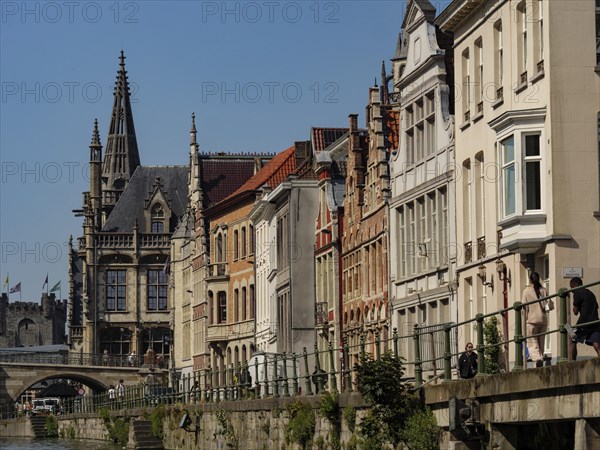 A street along a canal with gothic towers and historic houses under a clear blue sky, medieval facades with historic churches and buildings in a riverside town, Ghent, Belgium, Europe