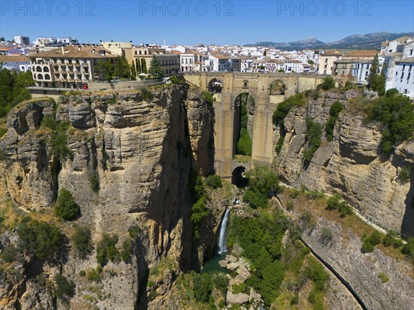 Breathtaking bridge over a deep gorge with waterfall and river, surrounded by rocks and buildings overlooking a sunny town and mountains, aerial view, waterfall, Ronda, Malaga, Andalucia, Spain, Europe