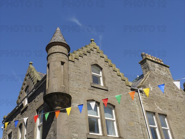 Colourful flags hang around a traditional building with a small tower and windows under a blue sky, grey houses with colourful flags in a seaside town, Lerwick, Scotland, United Kingdom, Europe