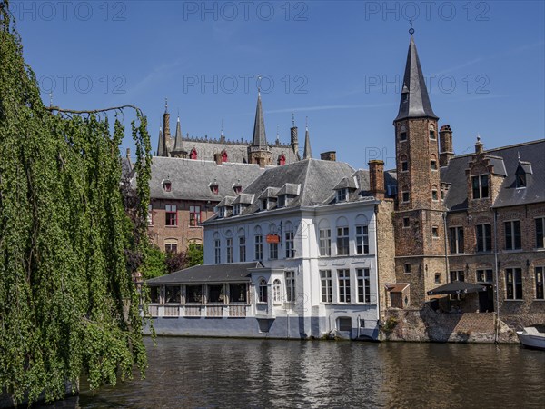 A building with a white facade and old brick towers by a river in sunny weather, historic houses and churches with towers by a river in Belgium, Bruges, Belgium, Europe
