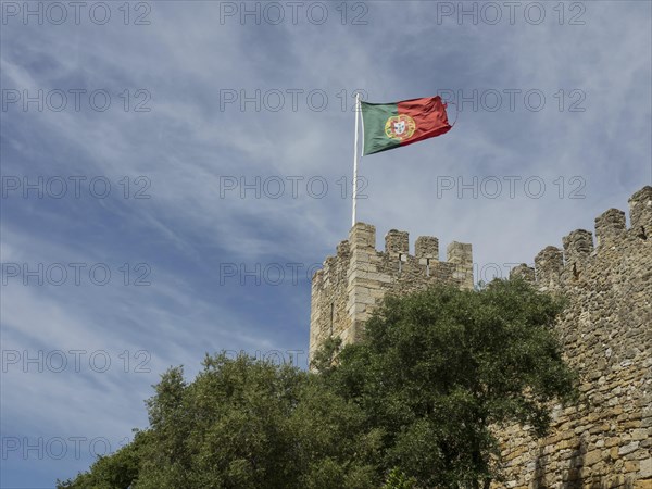 Part of a castle with the Portuguese flag on a watchtower spire, surrounded by green spaces and trees, Lisbon, Portugal, Europe