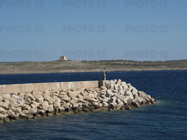 A breakwater of large stones in the sea with a rocky coastline in the background, the island of Gozo with historic houses, colourful balconies, magnificent churches and boats in the harbour, Gozo, Malta, Europe