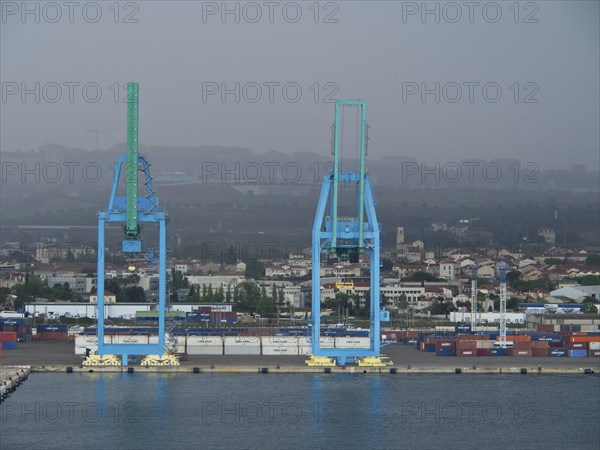 Two large blue harbour cranes in front of a cloudy industrial landscape, several containers in the harbour, Marseille on the Mediterranean Sea with the old cathedral and ships in the historic harbour, rocky coast on the Mediterranean Sea, marseille, france