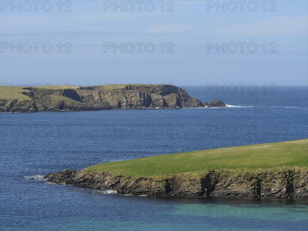 View of the blue sea with rocky cliffs and green grass in the foreground in calm weather, Green meadows by the sea with rocky shore and ruins, Lerwick, shetland