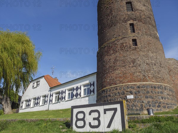 A stone tower and a house with the number 837 under a blue sky on a summer morning, rhine promenade of Rees with historic buildings green trees, rees, germany