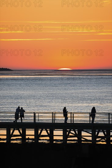 Silhouettes of several people on the pier Freilaufkanal, couple and single persons watch from a jetty the picturesque, golden, breathtakingly beautiful sunset over the sea, island Langluetjen II far away on the horizon, sun almost completely sunk, orange glowing cloudless sky, wide sea in soft colours, calm atmosphere, mouth of the Weser into the North Sea, Outer Weser, Bremerhaven, Land Bremen, Germany, Europe
