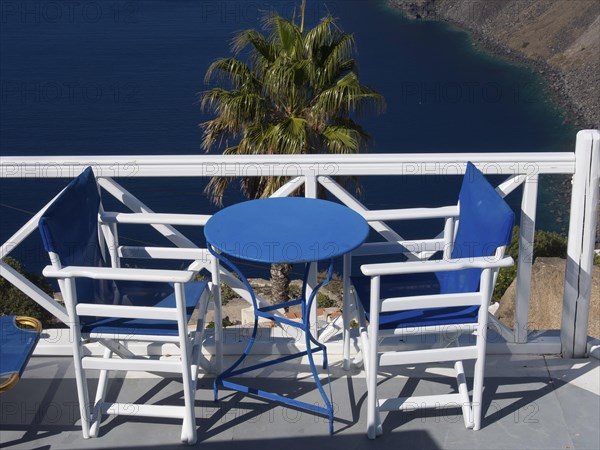 Two white chairs and blue table on a balcony with sea view and palm tree in the background, The volcanic island of Santorini with blue and white houses and churches, flowers and palm trees on a volcanic island, santorini, greece