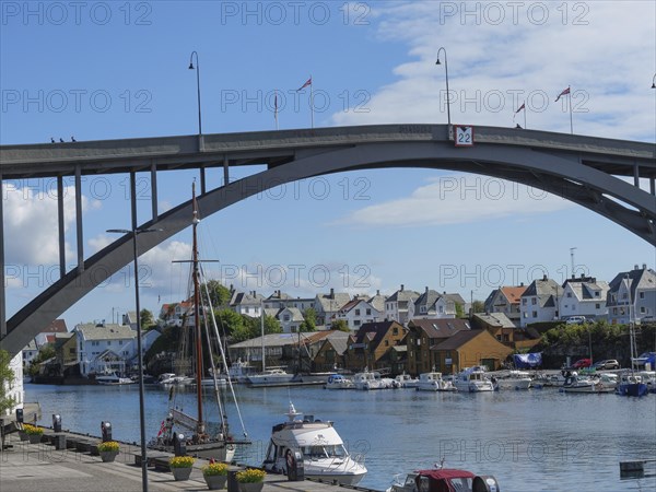 A large bridge over a waterway, including boats and a sailing ship, surrounded by urban houses and flags in the background, harbour in norway with boats, ships and many houses by the sea, Haugesund, Norway, Europe