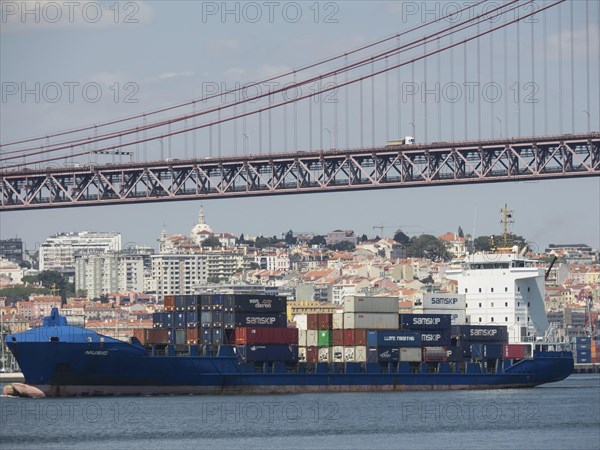 Large cargo ship loaded with containers under a bridge near the city on the waterfront, Lisbon, Portugal, Europe