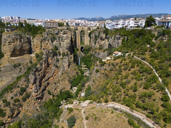 Impressive gorge with bridge, waterfall and town behind, surrounded by green landscape and mountains under bright sunshine, aerial view, Ronda, Malaga, Andalucia, Spain, Europe