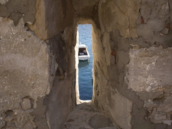 View through a narrow gap in an old stone wall to a boat in the sea, the old town of Dubrovnik with historic houses, churches, red roofs and fortress walls, dubrovnik, coratia
