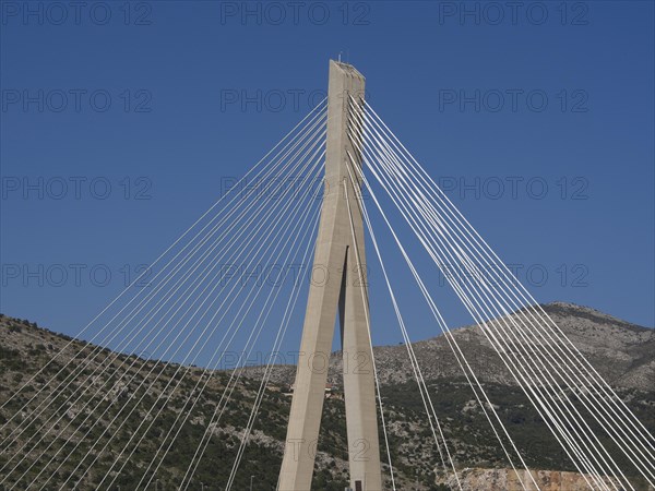 Modern suspension bridge with ropes in front of a clear blue sky and mountains, the old town of Dubrovnik with historic houses, churches, red roofs and fortress walls, dubrovnik, coratia