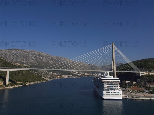 Suspension bridge over a river with a large cruise ship, surrounded by mountains and blue sky, the old town of Dubrovnik with historic houses, churches, red roofs and fortress walls, dubrovnik, coratia