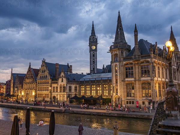 Gothic buildings with illuminated windows and people walking along a canal under a cloudy sky, historic buildings with towers and bridges by a river at blue hour, gent, belgium