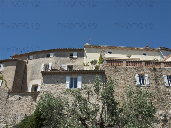 Stone buildings with windows and trees in the foreground under a blue sky, historic stone houses overgrown with greenery in Provence, Le Castellet, France, Europe