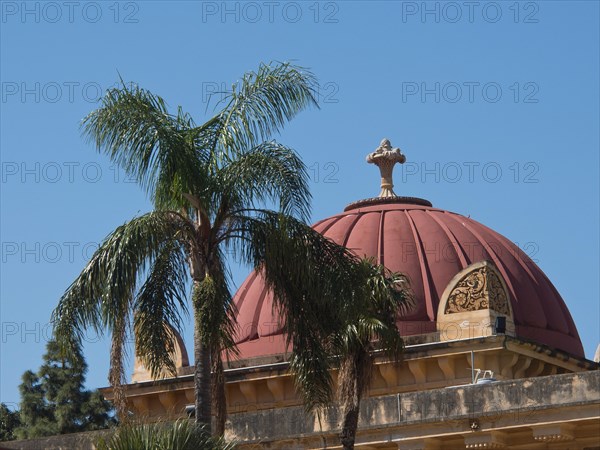 Historic building with red dome behind palm trees in front of a bright blue sky, palermo in sicily with an impressive cathedral, monuments and old houses with balconies, palm trees and flowers in italy, palermo, sicily