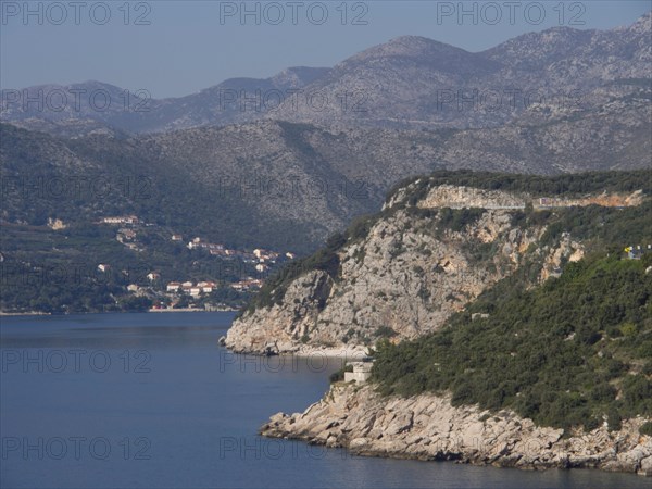 A rocky coastline flanked by green hills stretches into the calm sea, under a slightly cloudy sky, the old town of Dubrovnik with historic houses, churches, red roofs and fortress walls, dubrovnik, coratia