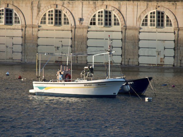 A boat in the harbour in front of a building with stone walls and many windows, small boats in the harbour in front of historic buildings in the evening light, Valetta, Malta, Europe