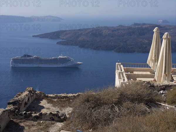A large cruise ship on the sea, rocky cliffs and comfortable sun loungers on a terrace, The volcanic island of Santorini with blue and white houses and churches, flowers and the deep blue sea, Santorini, Greece, Europe