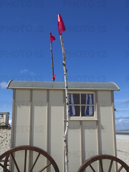 Side view of a beige changing cubicle with wooden wheels and two red flags under a blue sky, historic bathing trolleys on a sandy beach against a blue sky, Norderney, Germany, Europe