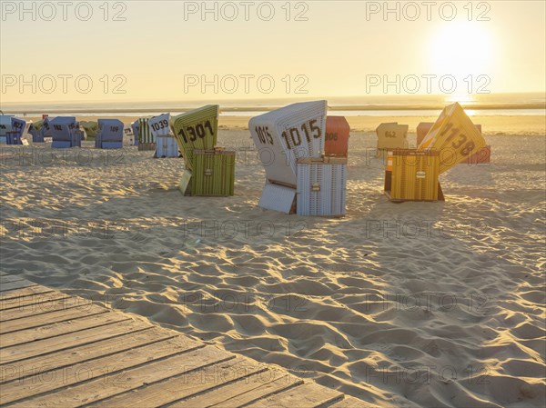 Several colourful beach chairs standing in order in the sand while the sun illuminates the horizon, a wooden path leads to the beach, setting sun on the North Sea beach with warm light over colourful beach chairs, langeoog, germany