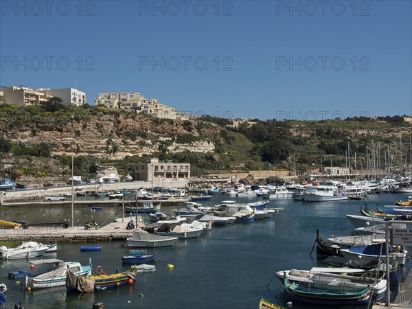 A quiet harbour with many sailing ships and fishing boats, surrounded by a hilly coastline and buildings under a blue sky, the island of Gozo with historic houses, colourful balconies, magnificent churches and boats in the harbour, Gozo, Malta, Europe