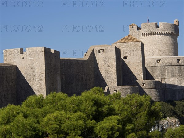 An imposing medieval fortress with high walls and towers rises into the blue sky, surrounded by trees, the old town of Dubrovnik with historic houses, churches, red roofs and fortress walls, dubrovnik, coratia