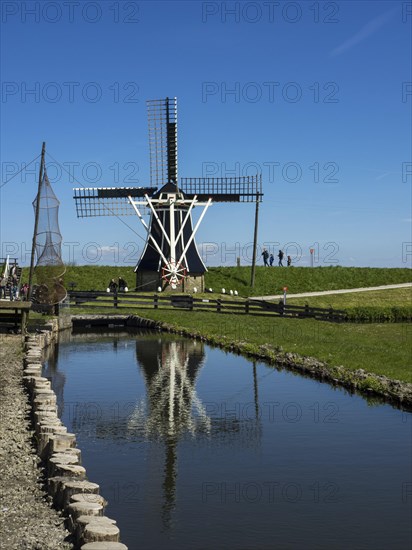 Large windmill with its reflection in a pond, surrounded by people, under a clear blue sky, Enkhuizen, Nirderlande