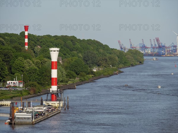Two red and white striped lighthouses on a river bank, in the background harbour facilities and industry, spring, houses and a red and white lighthouse on a green river bank, hamburg, germany
