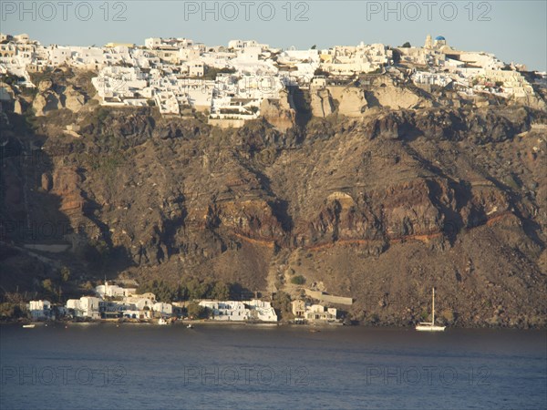 A cluster of white buildings on a cliff dramatically jutting out over the calm sea, brown volcanic island in the blue sea with white houses, santorini, greece